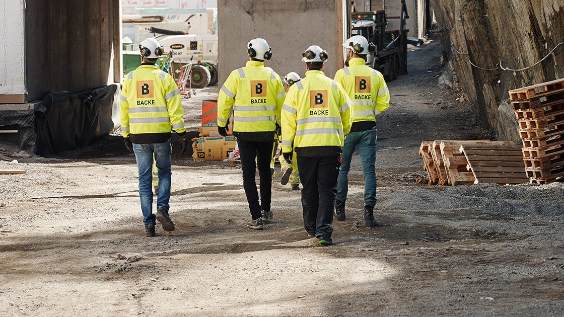 4 personer med ryggen til og Backe-logo på arbeidsklær går på en byggeplass